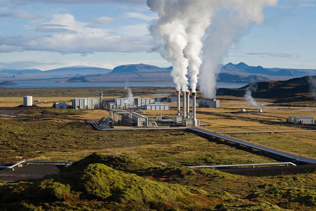 View of pipes in a valley by a lake with steam coming out the top.